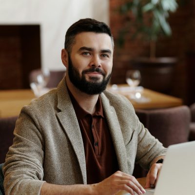 pexels-photo-4339954-4339954 Businessman with a beard and blazer using a laptop in a stylish cafeteria setting.